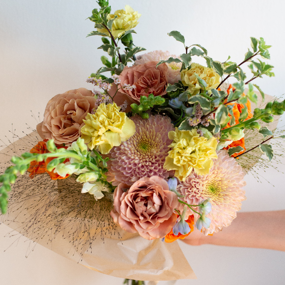 A hand holding a large, beautifully arranged floral bouquet featuring pale yellow carnations, dusty pink roses, chrysanthemums, and green foliage, wrapped in brown paper against a plain light background. Golden Hour Flower Studio in Oak Park, IL.