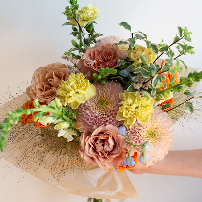 A hand holding a large, beautifully arranged floral bouquet featuring pale yellow carnations, dusty pink roses, chrysanthemums, and green foliage, wrapped in brown paper against a plain light background. Golden Hour Flower Studio in Oak Park, IL.